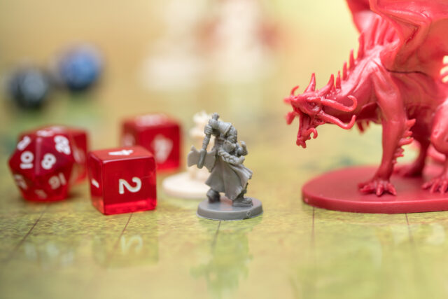 Photo with narrow depth of field, showing a tabletop roleplaying game. A grey plastic minature of a dwarf with a battle axe faces off against a large red minature of a dragon. Beside them, two glassy red six-sided dice and a similarly-coloured twenty-sided die can be seen.