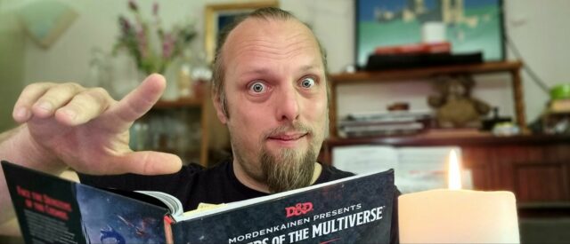 Dan gestures with a wave as he peers into the camera over the top of a copy of Monsters of the Multiverse, by candlelight.